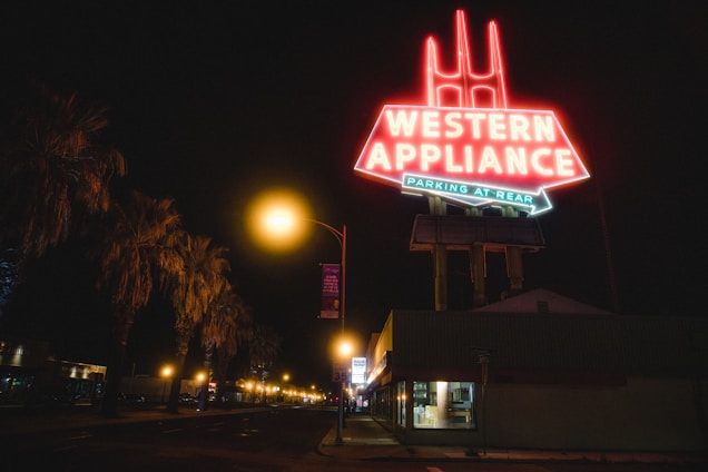 A brightly lit neon sign reading 'Western Appliance' dominates the night scene. The sign is elevated on tall supports and has additional text below indicating parking at the rear. The street is lined with palm trees, and the area is illuminated by streetlights along a seemingly quiet road. A building beneath the sign appears closed, with dimly lit windows.
