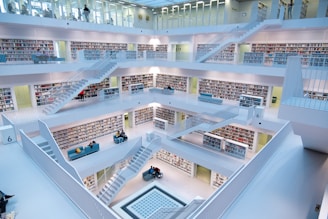 A modern, multi-level library features clean, white architectural design with multiple open floors connected by staircases. Each level has numerous bookshelves filled with books, and small seating areas with people reading and studying. The space is brightly lit with natural and artificial lighting providing a calm atmosphere.