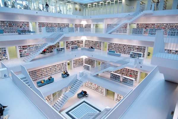 A modern, multi-level library features clean, white architectural design with multiple open floors connected by staircases. Each level has numerous bookshelves filled with books, and small seating areas with people reading and studying. The space is brightly lit with natural and artificial lighting providing a calm atmosphere.