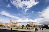 A bustling city square with colonial architecture, featuring a large cathedral and other historic buildings. People are walking, enjoying the open space, while the sky is partially cloudy with a vibrant blue hue.