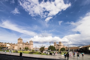 A bustling city square with colonial architecture, featuring a large cathedral and other historic buildings. People are walking, enjoying the open space, while the sky is partially cloudy with a vibrant blue hue.