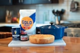 A kitchen scene featuring a bag of Gold Medal all-purpose flour, a container of Morton salt, a jar of Red Star active dry yeast, a loaf of bread, and a blue Dutch oven placed on a countertop. The background shows a blurred kitchen with a stove and utensils.