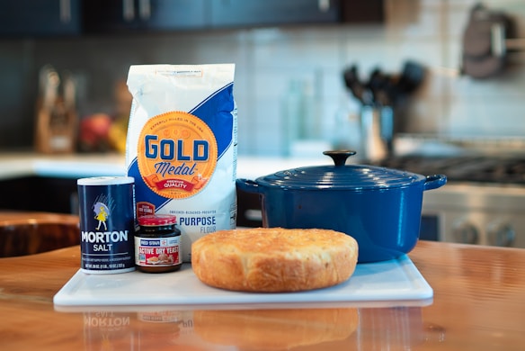 A kitchen scene featuring a bag of Gold Medal all-purpose flour, a container of Morton salt, a jar of Red Star active dry yeast, a loaf of bread, and a blue Dutch oven placed on a countertop. The background shows a blurred kitchen with a stove and utensils.