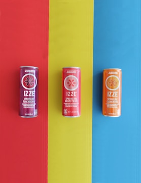 Three cans of IZZE sparkling drinks are arranged vertically on a colorful background. The cans are placed on a striking tri-color backdrop, with each can isolated on a separate color. The left can is on a red strip and features Sparkling Blackberry, the middle can on a yellow strip is Sparkling Grapefruit, and the right can on a blue strip is Sparkling Clementine. Each can displays its flavor name prominently with the brand's logo above.