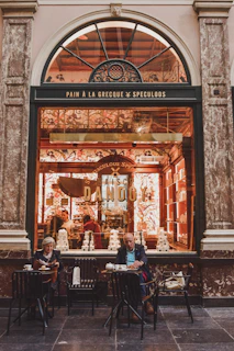 A charming storefront with an arched window displays a shop called Maison Dandoy. Intricately decorated with floral patterns, the interior is warmly lit, revealing piles of stacked boxes and a person working inside. In front of the shop, a man and a woman sit at small tables enjoying beverages and snacks.