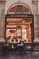 A charming storefront with an arched window displays a shop called Maison Dandoy. Intricately decorated with floral patterns, the interior is warmly lit, revealing piles of stacked boxes and a person working inside. In front of the shop, a man and a woman sit at small tables enjoying beverages and snacks.