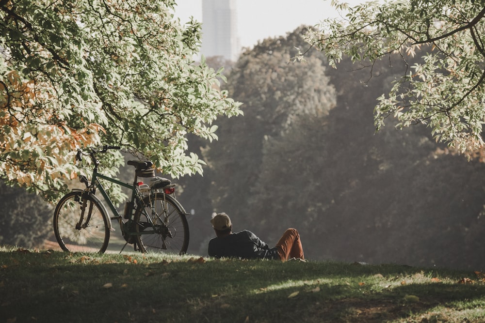 검은 재킷을 입은 남자가 낮에 검은 자전거 근처의 푸른 잔디밭에 앉아 있다