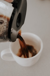 Hands pouring rich coffee from a French press into a clean white mug under morning light.