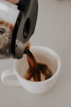 A stream of coffee is being poured from a French press into a white ceramic cup. The coffee is dark and rich, and the scene takes place on a light-colored countertop.