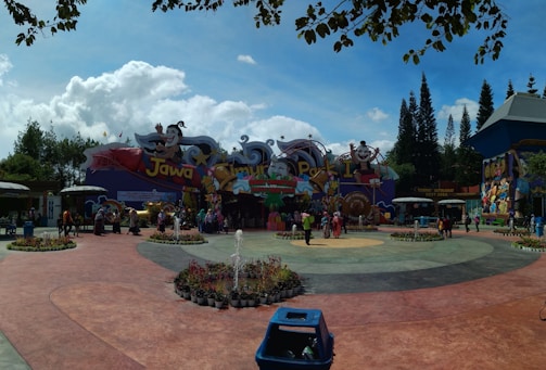 A vibrant amusement park entrance featuring colorful signage and cartoon characters. Visitors gather around a central plaza with landscaped greenery and a small fountain. The area is surrounded by tall trees and lively decorations.