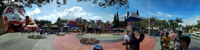 An amusement park entrance featuring vibrant and colorful facades, including a large structure resembling a face. People are gathered in groups, walking around, and taking photos. The sky is clear with a few clouds, and there are landscaped areas with flowers in the foreground.