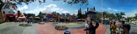 An amusement park entrance featuring vibrant and colorful facades, including a large structure resembling a face. People are gathered in groups, walking around, and taking photos. The sky is clear with a few clouds, and there are landscaped areas with flowers in the foreground.