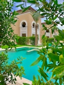 A luxurious building with beige walls and arched windows is surrounded by lush greenery and palm trees. In the foreground, a bright turquoise swimming pool is visible, bordered by well-maintained shrubs and trees.