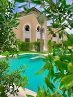 A luxurious building with beige walls and arched windows is surrounded by lush greenery and palm trees. In the foreground, a bright turquoise swimming pool is visible, bordered by well-maintained shrubs and trees.
