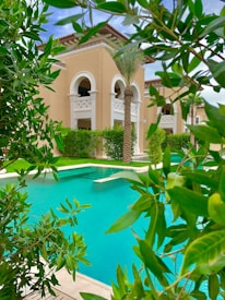 A luxurious building with beige walls and arched windows is surrounded by lush greenery and palm trees. In the foreground, a bright turquoise swimming pool is visible, bordered by well-maintained shrubs and trees.