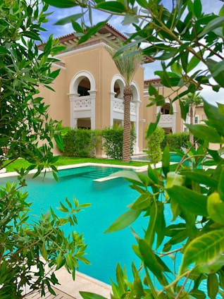 A luxurious building with beige walls and arched windows is surrounded by lush greenery and palm trees. In the foreground, a bright turquoise swimming pool is visible, bordered by well-maintained shrubs and trees.