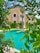 A luxurious building with beige walls and arched windows is surrounded by lush greenery and palm trees. In the foreground, a bright turquoise swimming pool is visible, bordered by well-maintained shrubs and trees.