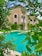 A luxurious building with beige walls and arched windows is surrounded by lush greenery and palm trees. In the foreground, a bright turquoise swimming pool is visible, bordered by well-maintained shrubs and trees.