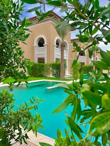 A luxurious building with beige walls and arched windows is surrounded by lush greenery and palm trees. In the foreground, a bright turquoise swimming pool is visible, bordered by well-maintained shrubs and trees.