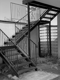 A metal staircase with ornate railings is situated outdoors. The stairs consist of two flights meeting at a square landing. The surrounding area includes a wall and some overgrown plants at the base.