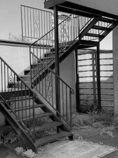 A metal staircase with ornate railings is situated outdoors. The stairs consist of two flights meeting at a square landing. The surrounding area includes a wall and some overgrown plants at the base.