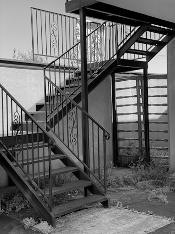 A metal staircase with ornate railings is situated outdoors. The stairs consist of two flights meeting at a square landing. The surrounding area includes a wall and some overgrown plants at the base.