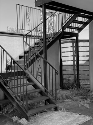 A metal staircase with ornate railings is situated outdoors. The stairs consist of two flights meeting at a square landing. The surrounding area includes a wall and some overgrown plants at the base.