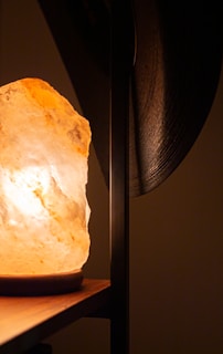 The image features a warmly lit Himalayan salt lamp on a wooden surface, casting a soft glow. The background is out of focus, with a dark vertical element partially visible to the right.