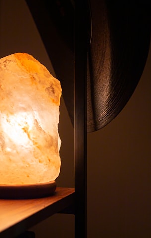 The image features a warmly lit Himalayan salt lamp on a wooden surface, casting a soft glow. The background is out of focus, with a dark vertical element partially visible to the right.
