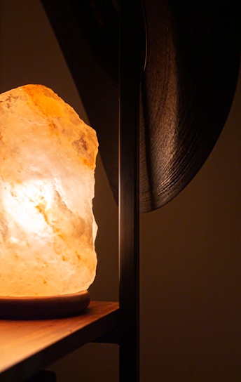 The image features a warmly lit Himalayan salt lamp on a wooden surface, casting a soft glow. The background is out of focus, with a dark vertical element partially visible to the right.