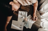 A person is leaning over a round wooden table, holding a pencil and making artistic sketches on paper. The sketches include an architectural drawing and color swatches. Sunlight filters through a curtain, casting soft shadows over the scene.