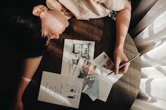 A person is leaning over a round wooden table, holding a pencil and making artistic sketches on paper. The sketches include an architectural drawing and color swatches. Sunlight filters through a curtain, casting soft shadows over the scene.