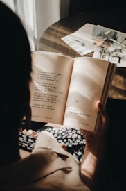 A person is holding an open book with text, sitting in a sunlit room. The sunlight creates a warm and cozy ambiance. On a table in the background, there are several magazines or papers.