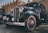 A classic vintage car featuring a sleek black body with red accents and whitewall tires is parked in what seems to be an outdoor area with rustic architecture in the background. Large round headlamps and a prominent front grille highlight its antique design.