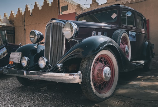 A classic vintage car featuring a sleek black body with red accents and whitewall tires is parked in what seems to be an outdoor area with rustic architecture in the background. Large round headlamps and a prominent front grille highlight its antique design.
