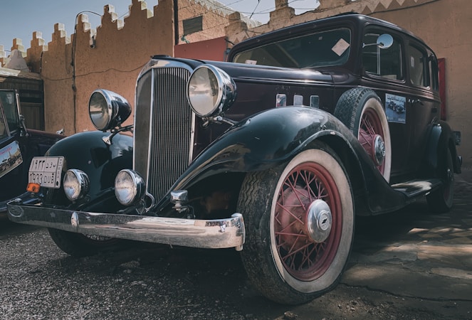 A classic vintage car featuring a sleek black body with red accents and whitewall tires is parked in what seems to be an outdoor area with rustic architecture in the background. Large round headlamps and a prominent front grille highlight its antique design.