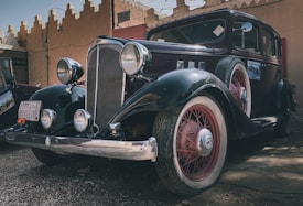 A classic vintage car featuring a sleek black body with red accents and whitewall tires is parked in what seems to be an outdoor area with rustic architecture in the background. Large round headlamps and a prominent front grille highlight its antique design.