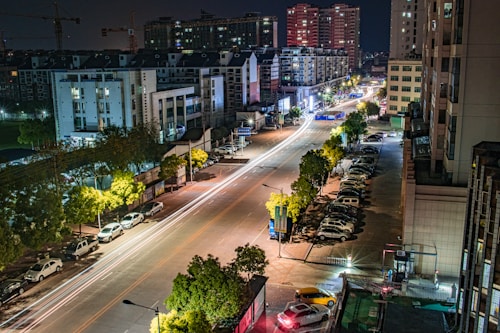 A nighttime cityscape depicting a well-lit urban street with light trails from moving vehicles. Tall buildings line both sides of the street, with parked cars visible along the roadside. Trees with illumination are scattered along the sidewalks, and more distant large buildings are seen in the background.