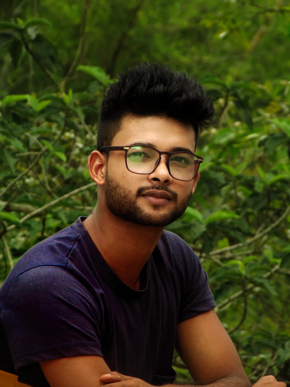 The image shows a young man with glasses and a beard, dressed in a blue shirt, seated on a bench surrounded by lush green trees. The man's gaze is directed off to the side, suggesting he may be lost in thought or simply enjoying his surroundings.