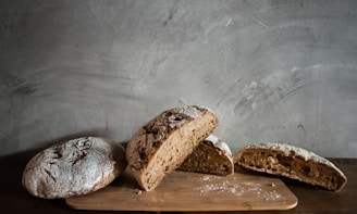 Several pieces of freshly baked bread are arranged on a wooden board against a textured gray wall. The bread is rustic, with a dusting of flour on top and a dense crumb structure.