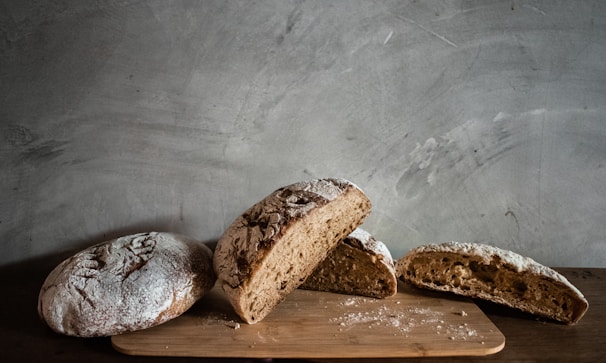 Several pieces of freshly baked bread are arranged on a wooden board against a textured gray wall. The bread is rustic, with a dusting of flour on top and a dense crumb structure.