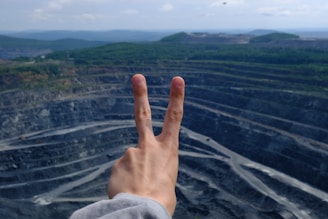 A hand showing a peace sign is held up against the backdrop of a large open-pit mine with tiered levels. The mine appears expansive and is surrounded by a landscape of green hills under a partly cloudy sky.