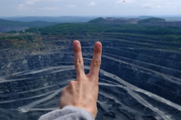 A hand showing a peace sign is held up against the backdrop of a large open-pit mine with tiered levels. The mine appears expansive and is surrounded by a landscape of green hills under a partly cloudy sky.