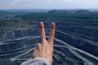 A hand showing a peace sign is held up against the backdrop of a large open-pit mine with tiered levels. The mine appears expansive and is surrounded by a landscape of green hills under a partly cloudy sky.