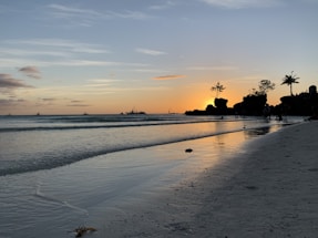 A tranquil golden beach at sunset with gentle waves and silhouetted villas in the distance.