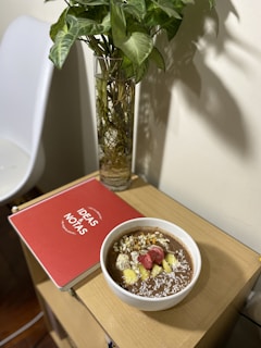 A wooden side table holds a red notebook labeled 'Ideas & Notas,' a bowl of what appears to be a healthy smoothie topped with sliced strawberries, bananas, seeds, and shredded coconut. Next to the table, there is a tall, clear vase containing a leafy plant with green foliage.