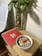 A wooden side table holds a red notebook labeled 'Ideas & Notas,' a bowl of what appears to be a healthy smoothie topped with sliced strawberries, bananas, seeds, and shredded coconut. Next to the table, there is a tall, clear vase containing a leafy plant with green foliage.