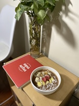 A wooden side table holds a red notebook labeled 'Ideas & Notas,' a bowl of what appears to be a healthy smoothie topped with sliced strawberries, bananas, seeds, and shredded coconut. Next to the table, there is a tall, clear vase containing a leafy plant with green foliage.