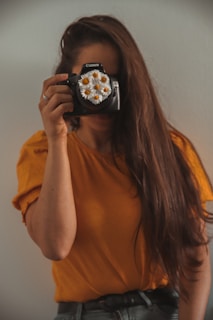 A person with long brown hair is holding a camera up to their face, obscuring it. The camera lens is decorated with white daisies. The person is wearing an orange shirt and standing against a light-colored background.