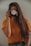A person with long brown hair is holding a camera up to their face, obscuring it. The camera lens is decorated with white daisies. The person is wearing an orange shirt and standing against a light-colored background.